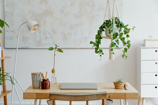 Stylish And Boho Home Interior Of Living Room With Wooden Desk, Laptop, White Lamp, Macrame Shelf And Desk Supplies. Design And Elegant Accessories. Modern Home Decor. Abstract Painting On The Wall. 