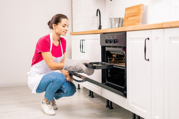 Young housewife squatting while putting tray with raw cookies into electric oven