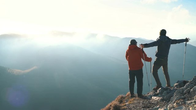 4K Two hikers standing on top of mountain stretching their hands in joy. Two men holding hiking poles facing extreme winds.