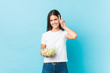 Young caucasian woman holding a salad showing victory sign and smiling broadly.