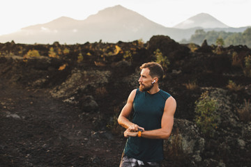 Naklejka premium Young hipster runner with beard using smart watch and listening music in wireless headphones before running outdoors.