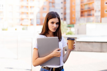 Portrait of smiling woman strolling through city street with silver laptop and takeaway coffee in hands