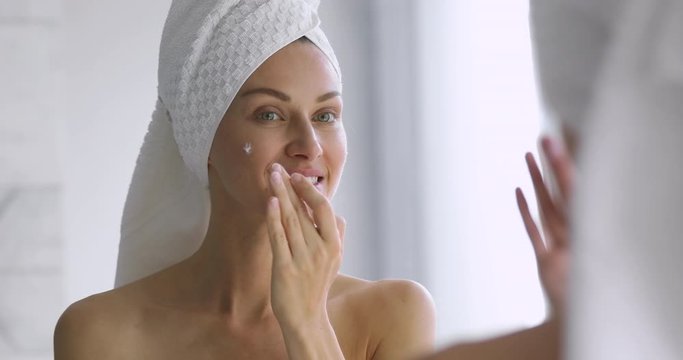 Funny woman apply skincare cream on face look in mirror