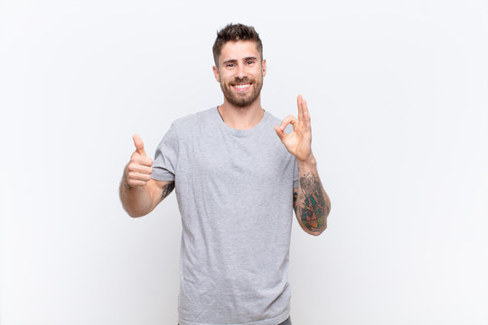 young handosme man feeling happy, amazed, satisfied and surprised, showing okay and thumbs up gestures, smiling against flat color wall