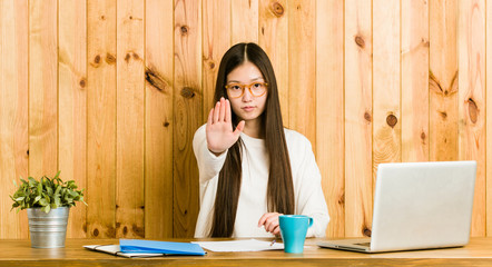 Young chinese woman studying on her desk standing with outstretched hand showing stop sign, preventing you.