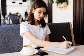 Young woman in gray dress sitting at table in cafe and writing in notebook. Student learning online.