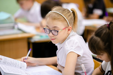 Smiling scholar girl sitting with other children in classroom and writing on textbook. Happy student doing homework at elementary school. Young schoolgirl feeling confident while writing on notebook