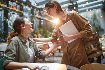 Side view portrait of two young women greeting each other joyfully during meeting in cafe on outdoor terrace decorated with lights
