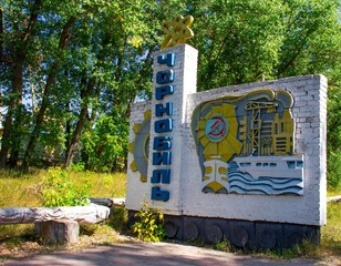 A sign with the name of Chernobyl written in Ukrainian. On top of the monument there is a yellow atom. On the side there is an industrial factory.Chernobyl entrance