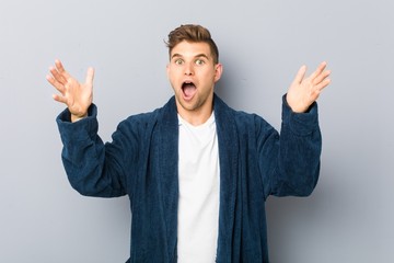 Young caucasian man wearing pajama receiving a pleasant surprise, excited and raising hands.