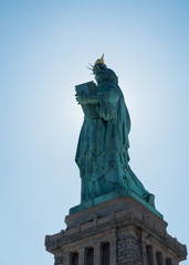 Obraz premium Back shot from statue of liberty with sun behind the statue and glowing edges. Torch, Pedestal and blue sky visible.