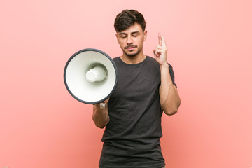 Young hispanic man holding a megaphone crossing fingers for having luck