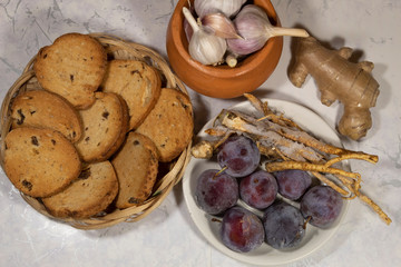 Natural products - on a wicker dish - bread toasts. On a saucer, frozen plums and parsley roots. Nearby is ginger root. In a clay pot - garlic.