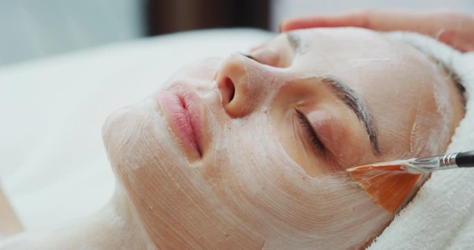 Close up of a professional cosmetologist applies facial clay mask on an young beautiful woman face before ultrasonic cleaning procedure for skin pores and deep moisturizing.