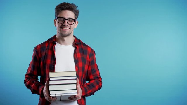 European student in red plaid shirt on blue background in studio holds stack of university books from library. Copy space. Guy smiles, he is happy to graduate.