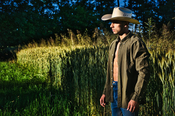 Portrait of sexy farmer or cowboy in hat with unbuttoned shirt on muscular torso, looking to a side, while standing next to hay field in countryside