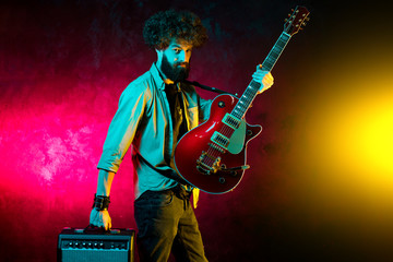 Portrait of hipster man with curly hair with red guitar in neon lights. Rock musician is playing electrical guitar.