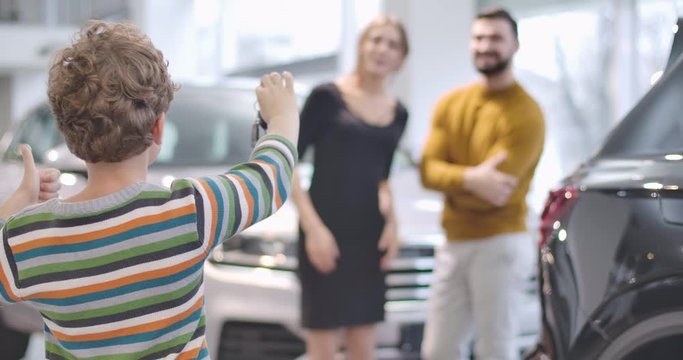 Back view of little Caucasian boy with curly hair bragging car keys and showing thumb up. Young parents smiling at the background. Successful family buying new vehicle. Cinema 4k footage ProRes HQ.