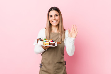 Young baker woman holding sweets smiling cheerful showing number five with fingers.