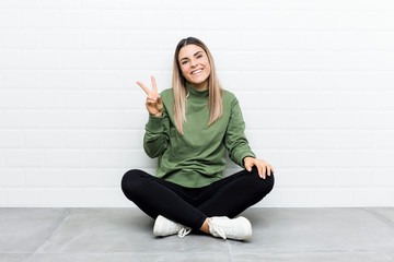 Young caucasian woman sitting on the floor joyful and carefree showing a peace symbol with fingers.