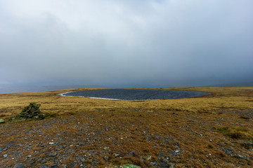 A view of a tarn (mountian lake) on a stony and grassy mountain plateau with a small cairn under a dark stormy sky