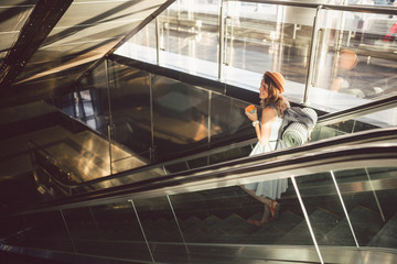 Portrait Smiling Woman Wearing Hat In Airport At Escalator. people traveling with hand luggage. Theme tourism and transport. Caucasian girl with coffee in airport terminal on an escalator