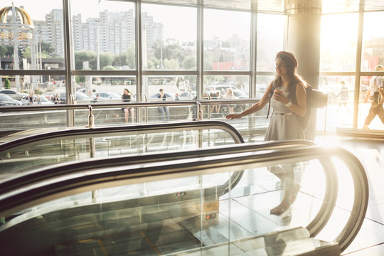 Portrait Smiling Woman Wearing Hat In Airport At Escalator. people traveling with hand luggage. Theme tourism and transport. Caucasian girl with coffee in airport terminal on an escalator