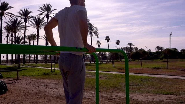 Slow motion beautiful backlight of a sportsman training on the bars of a park for calisthenics exercises and exercises with his own weight there are palm trees at the bottom of the image