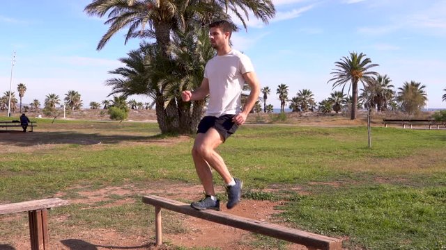 Slow motion, sportsman performing leg exercises, striding to climb the bench with one leg in a natural grass park