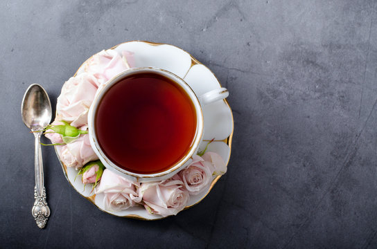 Tea time backdrop. Top-down still life with vintage white porcelain tea cup with gentle pink roses and nice tea spoon on a dark gray background.