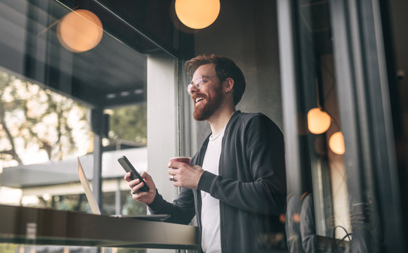 Delighted freelancer drinking and using smartphone in cafe