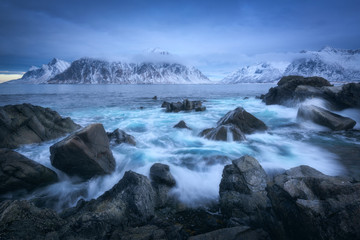 Fototapeta na wymiar Beach with stones in blurred waves, blue sky with low clouds and snowy mountains at dusk in winter. Sea coast in Lofoten islands, Norway. Dramatic landscape with sea, rocks in fog. Moody scenery