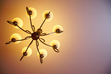 Lit wrought-iron vintage retro chandelier with eight shades hanging on the ceiling, on a light pink pastel background. Burning light bulbs. View from below
