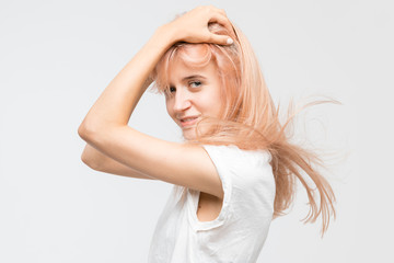 Naklejka premium Studio portrait of cute young attractive blonde girl in white t-shirt. Woman hold hands on head, look at camera and catty smile. Good-looking cheerful female. Youth and beauty concept.