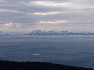 Winter landscape at sunrise. Silhouette of hills and trees in the foreground and snow capped mountain range of the European alps in the background.