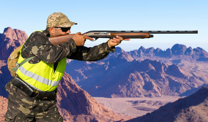 A hunter in camouflage and a gun in his hands in the early morning on top of a mountain stands...