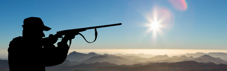 A hunter in camouflage and a gun in his hands in the early morning on top of a mountain stands...