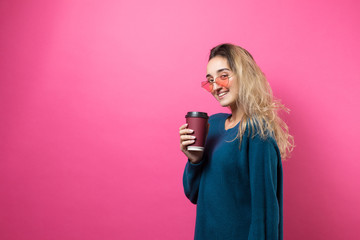 Glamor woman in glasses in a blue sweater with a drink of coffee on a pink background.