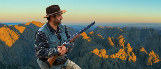 A hunter in camouflage and a gun in his hands in the early morning on top of a mountain stands looking for a target. Beautiful mountain landscape, the opening of the hunt.