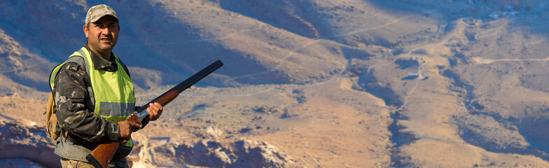 A hunter in camouflage and a gun in his hands in the early morning on top of a mountain stands...