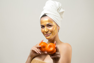 Beauty portrait of woman in white towel on head with gold nourishing mask on face. Skincare cleansing eco organic cosmetic spa relax concept. A girl stands with her back holding an orange mandarin.