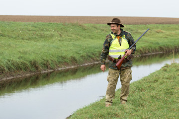 Naklejka premium Hunting period, autumn season open. A hunter with a gun in his hands in hunting clothes in the autumn forest in search of a trophy. A man stands with weapons and hunting dogs tracking down the game.