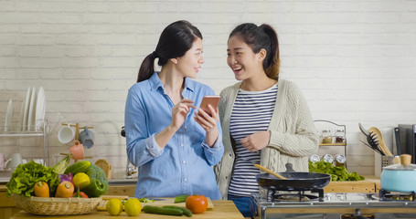 Teenage girl showing smartphone to best friend in kitchen in dormitory. young asian japanese women smiling happily while using cellphone in hands browsing online website on winter sale shopping