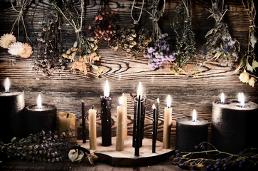 Black candles, herbs and flowers against wooden wall on witch table.