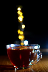 Glass Cup of hot tea on the table in the evening on a blurred dark background with yellow lights garland in the shape of steam rising up over the Cup