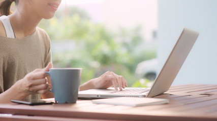Fototapeta premium Freelance Asian woman working at home, business female working on laptop and drinking coffee sitting on table in the garden in morning. Lifestyle women working at home concept.