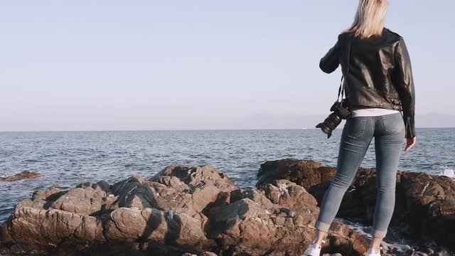 Woman in grunge style with DSLR camera on the shoulder standing on the rocks by the sea.