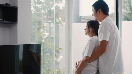 Young Asian Pregnant couple hug and holding belly talking with their child. Mom and Dad feeling happy smiling peaceful while take care baby, pregnancy near window in living room at home concept.