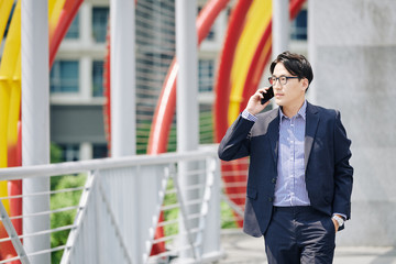 Fototapeta premium Serious Asian entrepreneur in glasses walking outdoors and talking on phone with business partner