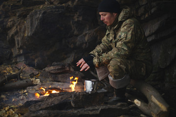 Fototapeta na wymiar Man in camouflage drinking tea and warms himself by the fire in bad weather. Extreme travel. Survival in the wild.
